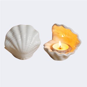 May include: Two white ceramic seashell candle holders. One is closed, the other open with a lit tea light candle inside. The candle holder has a textured, ridged surface and a warm, yellow glow from the candle.