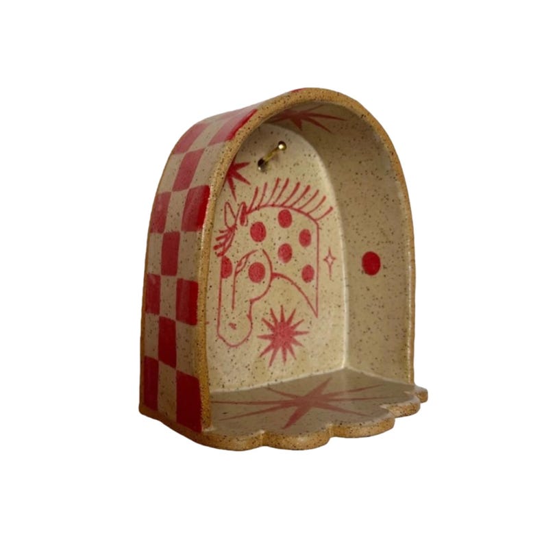 Pu&ograve; includere: Mensola in ceramica rossa e beige. Il lato sinistro presenta un motivo a scacchi rossi, mentre l'interno mostra un'illustrazione di cavallo rosso con dettagli a stella e puntini. La mensola ha una base smerlata e un piccolo gancio in ottone.