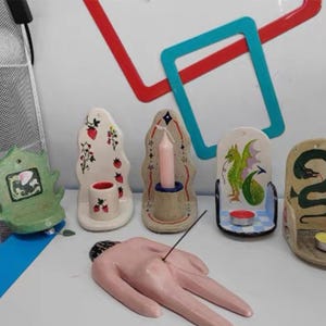 Pu&ograve; includere: Una collezione di portacandele e porta incenso in ceramica. I portacandele presentano vari disegni, tra cui fragole, un drago e un serpente. Il porta incenso ha la forma di una mano. La tavolozza dei colori comprende bianco, rosa, verde e blu.