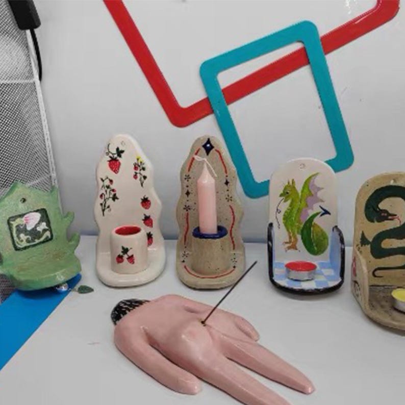 Pu&ograve; includere: Una collezione di portacandele e porta incenso in ceramica. I portacandele presentano vari disegni, tra cui fragole, un drago e un serpente. Il porta incenso ha la forma di una mano. La tavolozza dei colori comprende bianco, rosa, verde e blu.