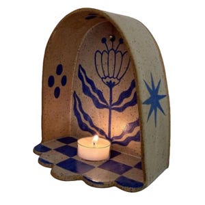 Pu&ograve; includere: Un portacandele in ceramica con finitura beige macchiettata, con disegni floreali e a stella blu. L'interno presenta un motivo a scacchi blu e bianco e all'interno &egrave; presente una candela a lumino accesa.