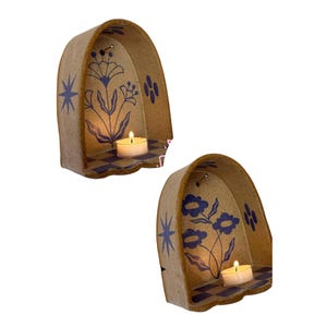 Pu&ograve; includere: Due portacandele in ceramica con base beige e disegni floreali blu. Ogni portacandele ha una piccola candela accesa all'interno. I portacandele hanno una forma ad arco curvo con un motivo a scacchi sulla base e disegni di stelle e fiori.