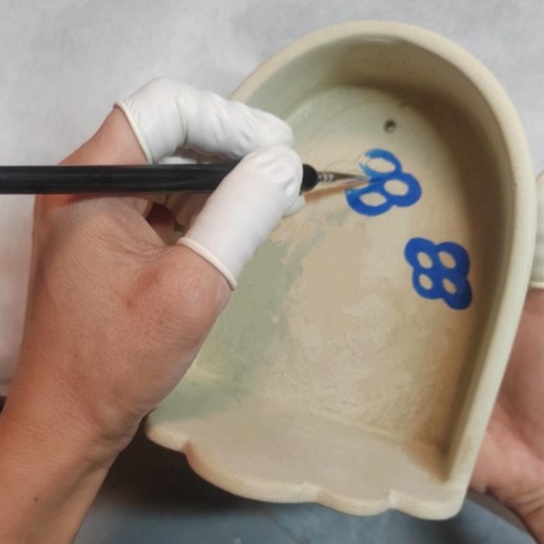 Pu&ograve; includere: Una persona sta dipingendo disegni floreali blu su un pezzo di ceramica beige. Il pezzo di ceramica ha un bordo smerlato e una parte superiore curva. La persona indossa guanti bianchi e usa un pennello sottile per applicare la vernice.