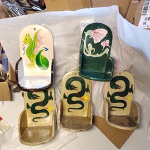 Pu&ograve; includere: Una collezione di portabicchieri da parete decorativi dipinti a mano. I portabicchieri presentano vari disegni, tra cui draghi e motivi floreali. La tavolozza dei colori include verde, rosa, giallo e crema. Ogni portabicchieri ha un piccolo ripiano curvo.