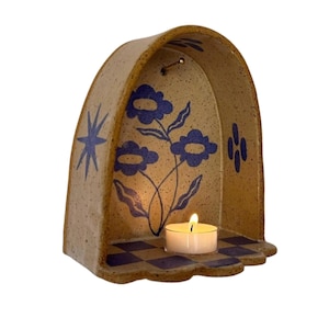 Pu&ograve; includere: Un portacandele in ceramica con finitura beige e disegni floreali e geometrici blu. La struttura ad arco presenta un piccolo ripiano con una candela tealight accesa. La base ha un motivo a scacchiera.