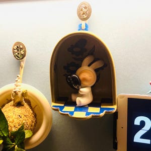 Pu&ograve; includere: Una stravagante statuina di coniglio in ceramica che tiene un cuore nero, all'interno di una mensola decorativa gialla e blu. La mensola &egrave; montata su una parete azzurra, con un motivo floreale sul retro.