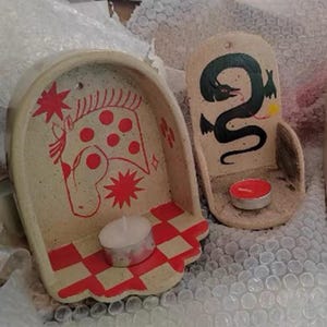 Pu&ograve; includere: Due portacandele in ceramica. Uno presenta un disegno di cavallo rosso con stelle, l'altro un serpente nero. Ognuno ha una piccola candela. Le basi hanno un motivo a scacchi rossi e bianchi.