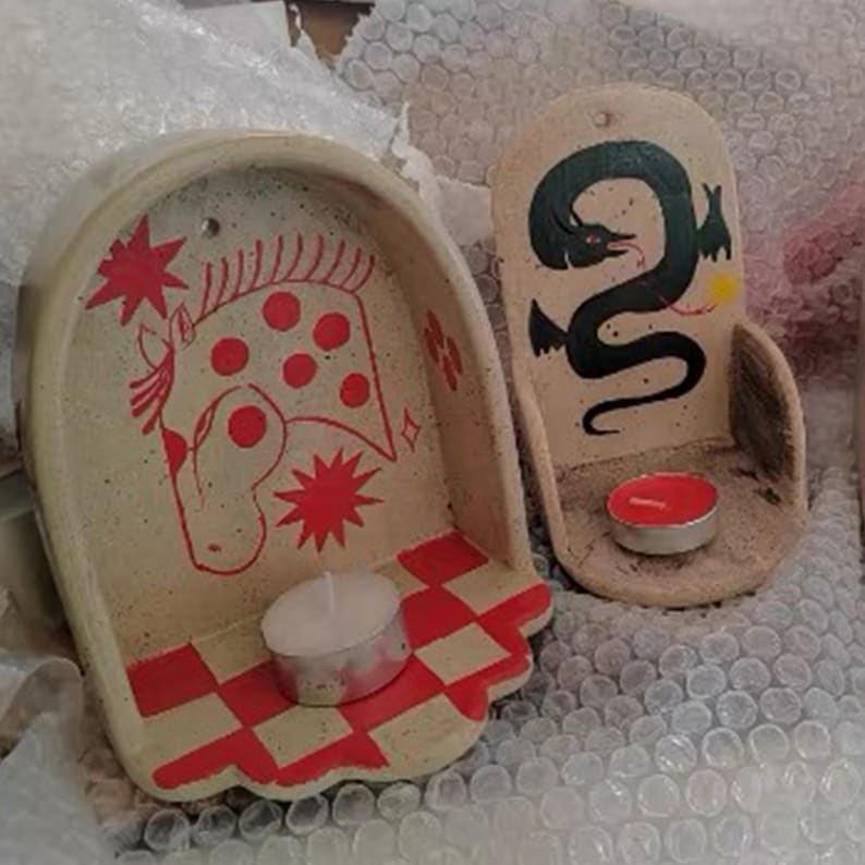 Pu&ograve; includere: Due portacandele in ceramica. Uno presenta un disegno di cavallo rosso con stelle, l'altro un serpente nero. Ognuno ha una piccola candela. Le basi hanno un motivo a scacchi rossi e bianchi.