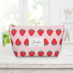 Op de afbeelding: Een roze toilettas met rits en een herhalend patroon van rode aardbeien op een roze achtergrond. De tas heeft een wit label met de naam "Jennika" in zwarte cursieve letters.