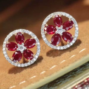 May include: A pair of floral-shaped stud earrings. Each earring features five oval-cut, deep red gemstones arranged around a central white gemstone. The gemstones are set within a halo of small, clear stones, creating a sparkling effect. The earrings are set in a gold-colored metal.