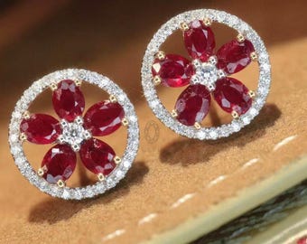 May include: A pair of floral stud earrings. Each earring has five oval, deep red gemstones around a central white gemstone. The gemstones are set within a halo of small, clear stones, creating a sparkling effect. The earrings are set in a gold-coloured metal.