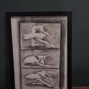 May include: A framed pencil drawing of three ballet dancers in motion. Each dancer is shown in a separate rectangular panel, displaying different poses. The artwork is in grayscale, with a black frame, and is on a wooden surface.
