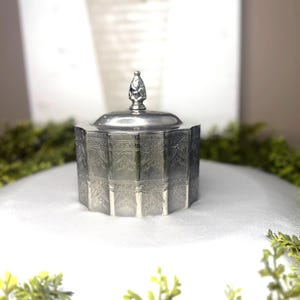 May include: A silver-colored, octagonal trinket box with a lid and a decorative finial. The box features embossed floral patterns on its sides. The background includes greenery and a blurred image of a white vase with flowers.