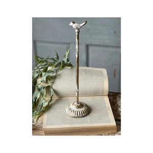 May include: A distressed, off-white and gold-colored bird-topped stand. The bird sits atop a slender pole, which is attached to a decorative, round base. The stand is placed on an open book with aged pages.