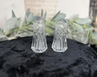 May include: A pair of clear glass salt and pepper shakers featuring a detailed geometric cut crystal pattern. Each shaker has a rounded top and a flared base, with vertical and crisscross etched lines reflecting light across the transparent surface.