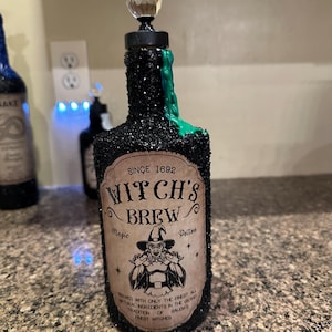 Puede incluir: Una botella negra cubierta de purpurina con un tapón de cristal y goteo verde. La etiqueta dice "Witch's Brew" con una ilustración de bruja. La botella es una botella de poción, perfecta para la decoración de Halloween.