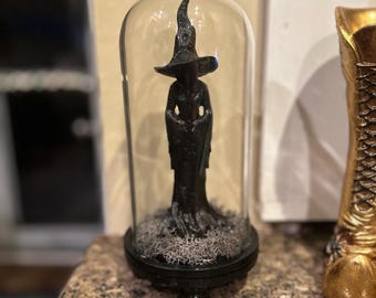 May include: A black witch figurine inside a glass cloche with a crystal-like topper. The witch is wearing a pointed hat and a long robe. The base is black and contains dried moss. A gold boot is visible in the background.