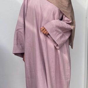 May include: A long-sleeved, light purple dress with a loose fit. The dress has a simple design with wide sleeves and a modest neckline. A brown head covering is draped over the head and shoulders. The dress appears to be made of a lightweight fabric.