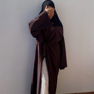以下が含まれることがあります： 長いダークブラウンのローブに、リボンベルトと長袖が付いています。ローブは白いドレスの上に着用されており、その人は黒いスカーフを着用しています。