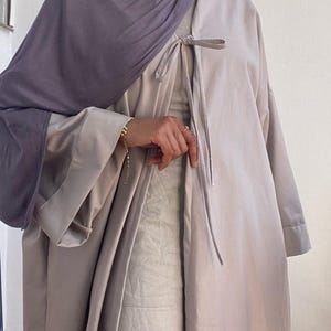 以下が含まれることがあります： 首元に結び目のある留め具が付いた、薄いグレーの長袖アバヤ。アバヤは薄いベージュのインナーの上に着用されています。紫色のスカーフが頭と肩にかけられています。