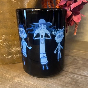 May include: Black ceramic mug with a white design of three cartoon characters with large eyes. The characters are a woman with a hat, a girl, and a boy. The characters are all wearing white clothing and have blue eyes.
