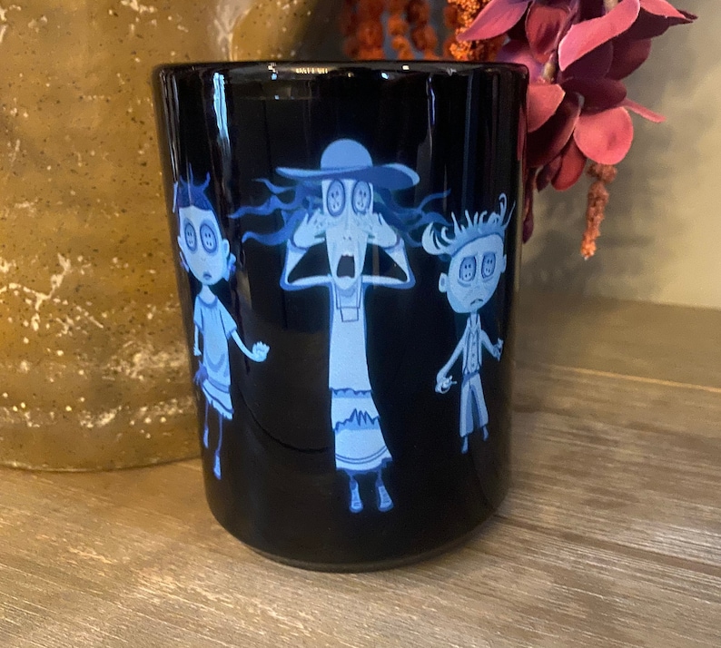 May include: Black ceramic mug with a white design of three cartoon characters with large eyes. The characters are a woman with a hat, a girl, and a boy. The characters are all wearing white clothing and have blue eyes.
