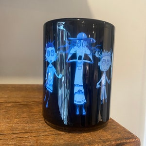 May include: A black ceramic mug with a white design of three cartoon characters with button eyes. The characters are a woman with a hat, a man, and a girl. The mug is on a wooden surface.