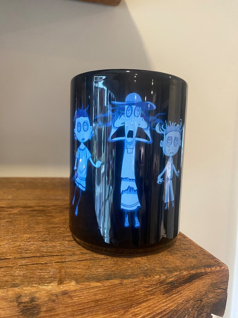 May include: A black ceramic mug with a white design of three cartoon characters with button eyes. The characters are a woman with a hat, a man, and a girl. The mug is on a wooden surface.