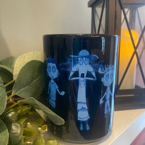 May include: A black ceramic mug with a blue and white design of three cartoon characters with button eyes. The characters are a woman with a hat, a boy, and a girl. The mug is on a white surface.