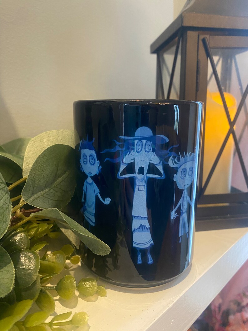 May include: A black ceramic mug with a blue and white design of three cartoon characters with button eyes. The characters are a woman with a hat, a boy, and a girl. The mug is on a white surface.