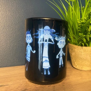 May include: Black ceramic mug with a white design of three cartoon characters with large eyes. The characters are a woman with a hat, a girl with short hair, and a boy with short hair. The characters are all looking surprised.