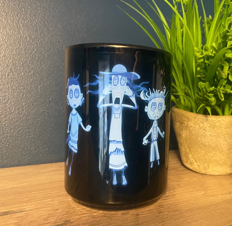 May include: Black ceramic mug with a white design of three cartoon characters with large eyes. The characters are a woman with a hat, a girl with short hair, and a boy with short hair. The characters are all looking surprised.