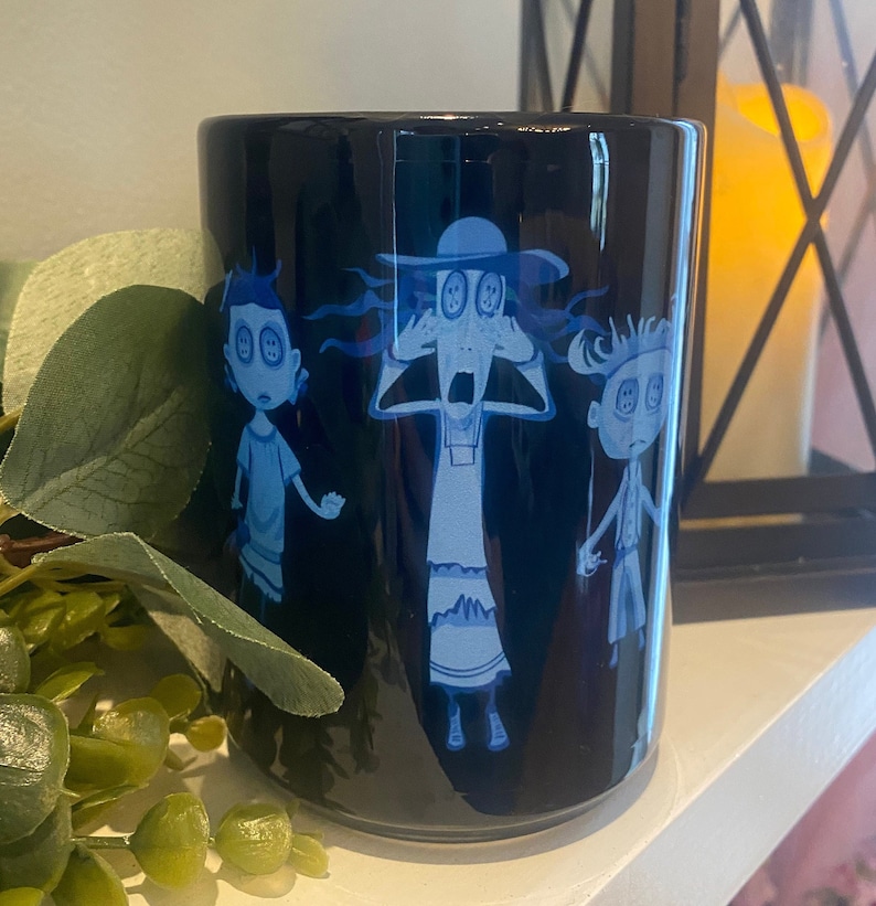May include: Black ceramic mug with a blue and white illustration of three cartoon characters with large eyes. The characters are a woman with a hat, a man, and a woman. The woman with the hat is covering her eyes with her hands and has a surprised expression. The man is looking to the left and the woman is looking to the right.