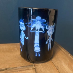 May include: A black ceramic mug with a white design of three cartoon characters with large eyes. The characters are a woman with a hat, a girl, and a boy. The mug is on a wooden surface.