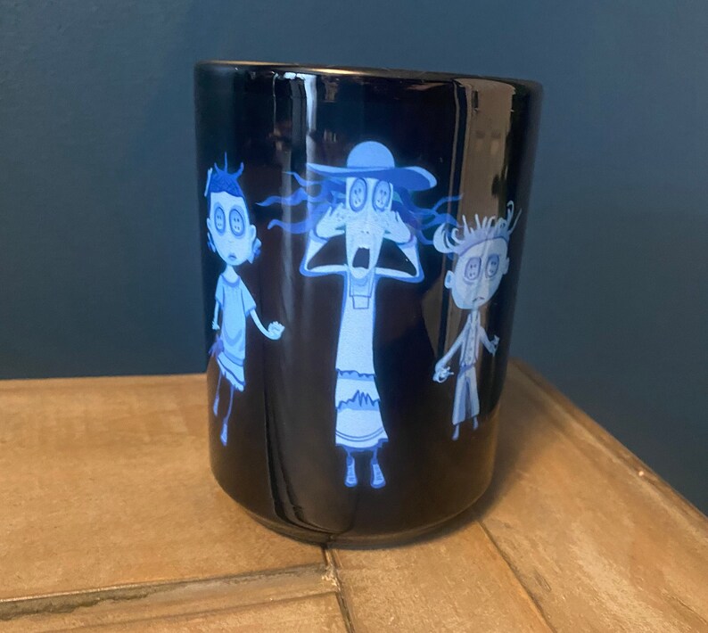 May include: A black ceramic mug with a white design of three cartoon characters with large eyes. The characters are a woman with a hat, a girl, and a boy. The mug is on a wooden surface.