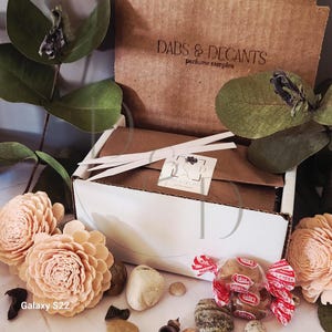 May include: A white box with a brown lid, containing perfume samples, is surrounded by decorative elements. The box is labeled "DABS & DECANTS perfume samples." Decorative peach-colored flowers, small stones, and wrapped candies are also present.