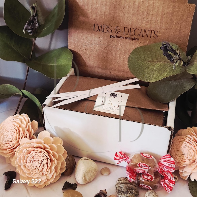 May include: A white box with a brown lid, containing perfume samples, is surrounded by decorative elements. The box is labeled "DABS & DECANTS perfume samples." Decorative peach-colored flowers, small stones, and wrapped candies are also present.