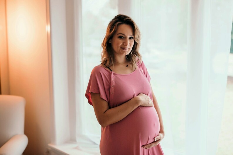 Plus Size pregnant mom wearing a light pink hospital delivery gown to deliver her baby. Fabric is soft and stretchy with snaps at shoulder for breastfeeding and down back for epidural. Long enough to cover knees. Favorite baby shower gift from Mom.