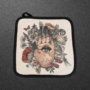 May include: A square pot holder with a black border and a hanging loop. The center features a detailed illustration of a hand with an eye in the palm, surrounded by various botanical elements including mushrooms, berries, ferns, and flowers.