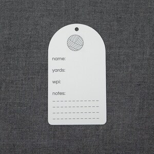 May include: A white, arch-shaped yarn inventory tag featuring a small hole at the top. The tag displays a circular yarn ball icon and printed text including name, yards, wpi, and notes, followed by four dashed lines for writing information.