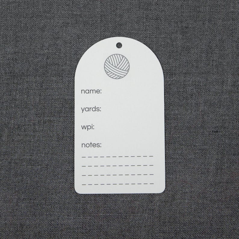 May include: A white, arch-shaped yarn inventory tag featuring a small hole at the top. The tag displays a circular yarn ball icon and printed text including name, yards, wpi, and notes, followed by four dashed lines for writing information.