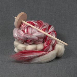 May include: A wooden drop spindle resting on a coiled bundle of unspun wool fiber. The fiber features a blend of solid white and variegated red and white sections, arranged in a thick, braided texture with loose, soft ends extending outward.
