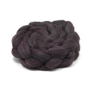 May include: A thick, dark purple wool roving braided into a dense, circular coil. The fiber has a soft, fuzzy texture with visible strands and deep, rich tones throughout the intertwined structure of the coiled material.