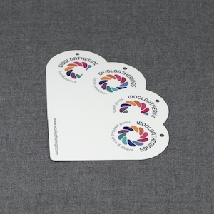May include: Four white, rounded-top rectangular tags stacked in a fan shape. Each tag features a circular logo with colorful segments and the text Woolgathering, hand painted, and yarn & fibers. The website www.woolgatherings.com is printed on the side.