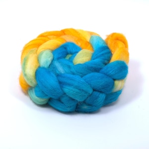 May include: A coiled bundle of roving wool fiber featuring a vibrant gradient of colors. The texture is soft and fluffy, transitioning from bright yellow and orange tones into shades of light green, turquoise, and deep blue in a braided pattern.