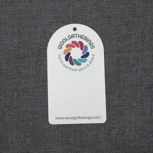 May include: A white, arched, rectangular tag featuring a circular logo with multi-colored petal shapes. The text reads WOOLGATHERINGS, hand painted yarn & fibers, and the website address www.woolgatherings.com. There is a small hole at the top center.