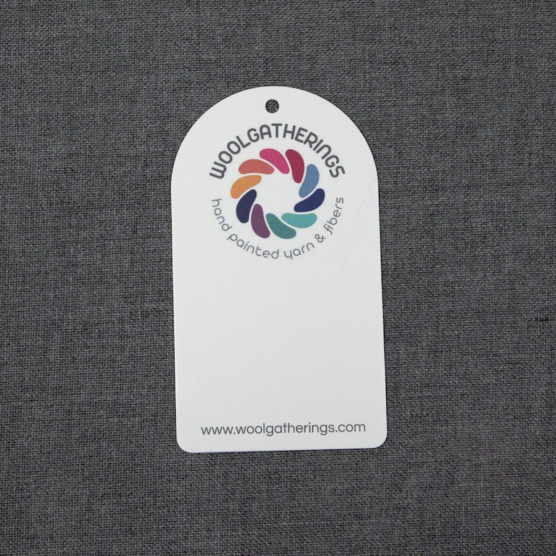 May include: A white, arched, rectangular tag featuring a circular logo with multi-colored petal shapes. The text reads WOOLGATHERINGS, hand painted yarn & fibers, and the website address www.woolgatherings.com. There is a small hole at the top center.