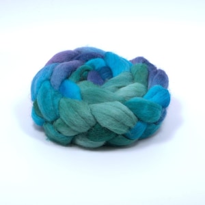 May include: A thick, braided bundle of soft wool roving featuring a gradient of colors ranging from deep navy blue and royal blue to vibrant turquoise, emerald green, and soft seafoam green, arranged in a circular, coiled, and textured form.