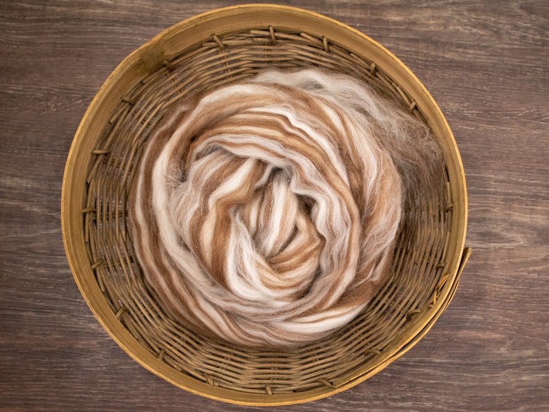 May include: A coiled bundle of soft, unspun wool roving featuring a marbled blend of white, cream, and various shades of brown fibers. The wool is contained within a round, woven wooden basket with a light brown finish and visible slats.