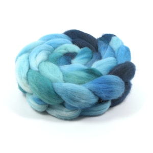May include: A thick, braided bundle of soft wool roving featuring a gradient of colors ranging from pale sky blue and turquoise to deep navy blue. The fiber is coiled into a circular shape, showcasing a textured, fluffy appearance throughout.